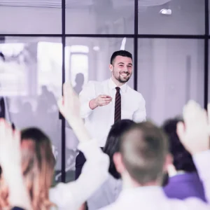 Man at the front of a seated crowd points at someone raising their hand, signifying cpd-accredited webinars and courses