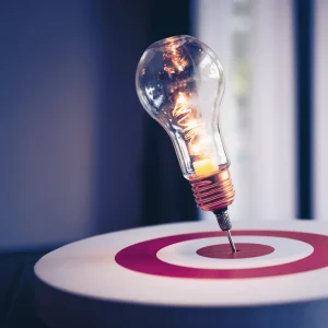 A lit light bulb is stuck at the centre of a target, signifying solving systemic problems