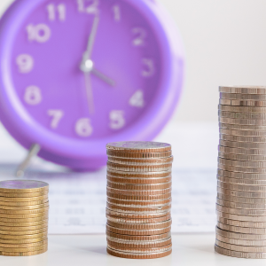 Three increasing stacks of coins sit in front of a purple clock. To signify how transaction management can save time and increase cost efficiencies.